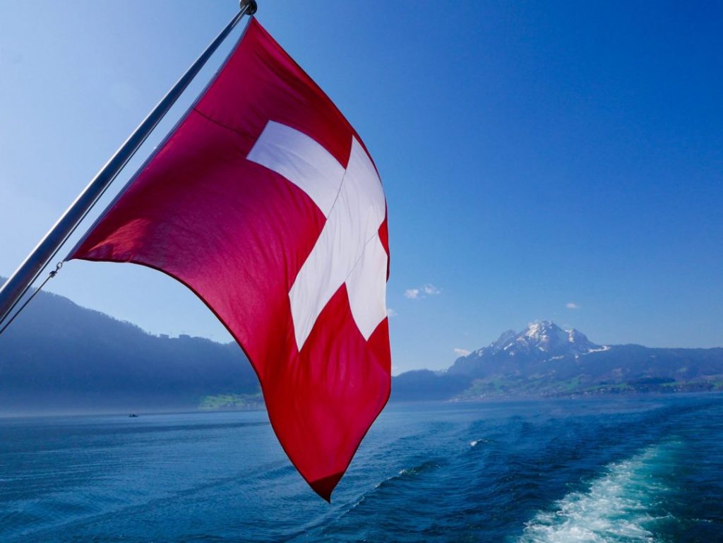 the swiss flag from the back of a boat with mountains in the background. Switzerland is one of the best solo female travel destinations!