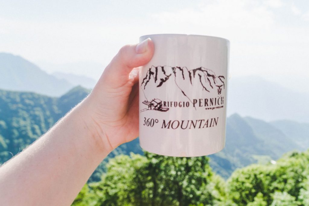 Hand holding a mug from Rifugio Pernici in front of a mountainous background in Valle di Ledro, Italy
