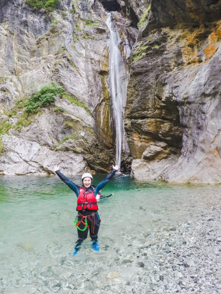 Addie standing confidently with her hands over her head in front of a tall, thin waterfall
