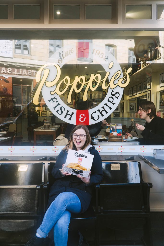 addie sitting on a bench in front of poppie's fish and chips holding a box of fish and chips and looking super excited - a perfect place to stop for food on a girls weekend in london
