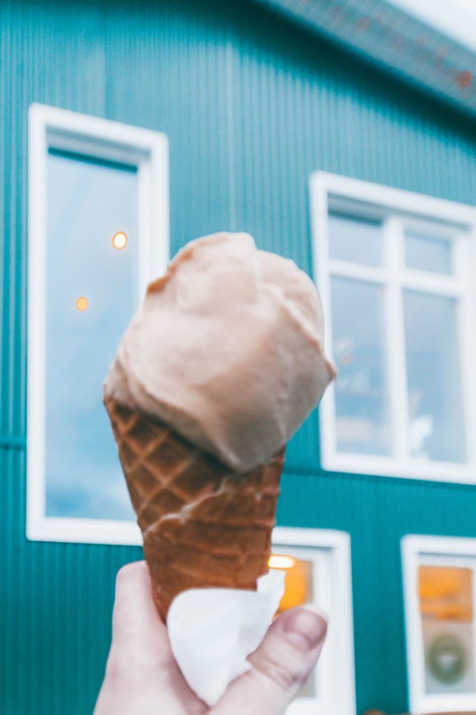 Salted carmel ice cream in a cone, held up in front of the green building at Efstidalur Dairy Farm
