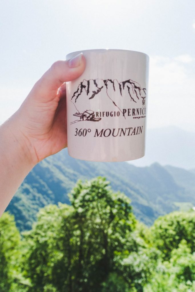 A hand holding a mug from Rifugio Pernici in front of a mountainous background