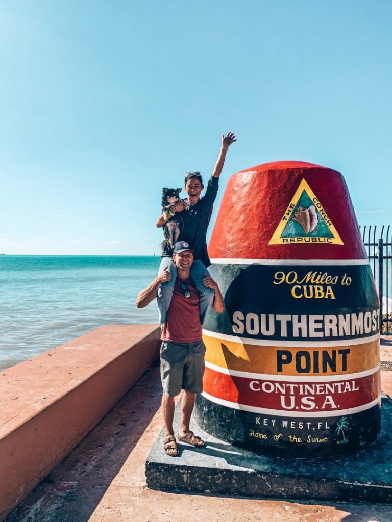 a woman holding a small dog and sitting on a man's shoulders in front of the southernmost point marker in Key West, Florida