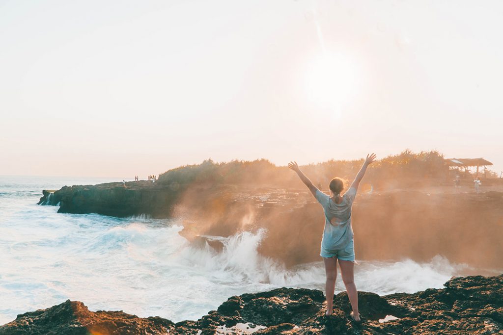 addie standing with her hands in the air as mist comes up from devil's tear on nusa lembongan - nusa islands guide