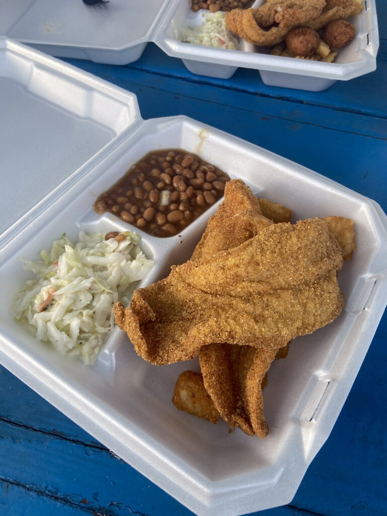 Takeout container with fried catfish, baked beans, and coleslaw on a blue table. Another similar container is partially visible in the background. E'Town River Restaurant is perfect as part of your Shawnee National Forest itinerary.