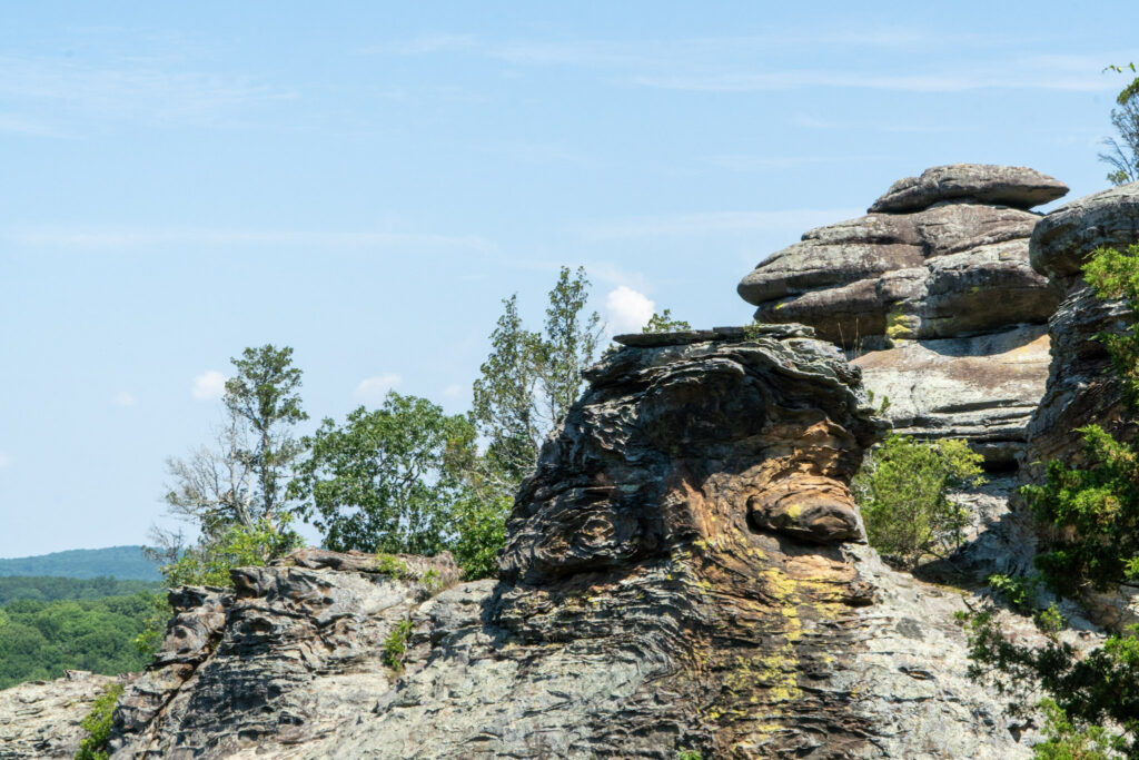 A scenic view of weathered, layered rock formations with patches of green vegetation, set against a backdrop of blue sky and distant green hills. The rock formations appear rugged and natural.