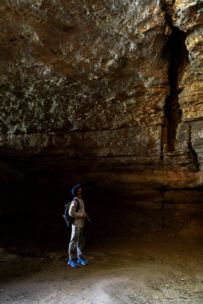 A person in hiking attire and a baseball hat, holding a camera, stands inside a large, dimly lit cave within Cave-in-Rock State Park. They appear to be looking up at the high, textured rock ceiling. The ground is uneven, and the cave walls are rugged with shades of brown and ochre.