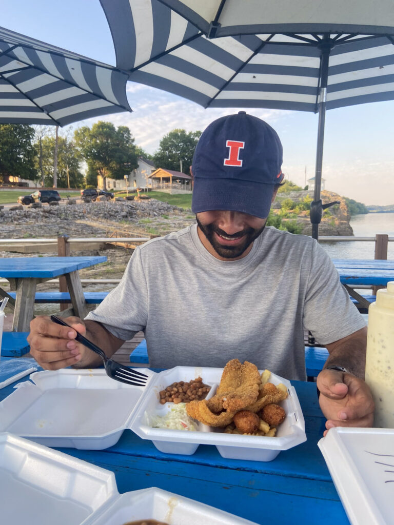 A person in a cap sits at an outdoor table with a plate of food, including fried items, beans, and coleslaw, under a striped umbrella on a sunny day.