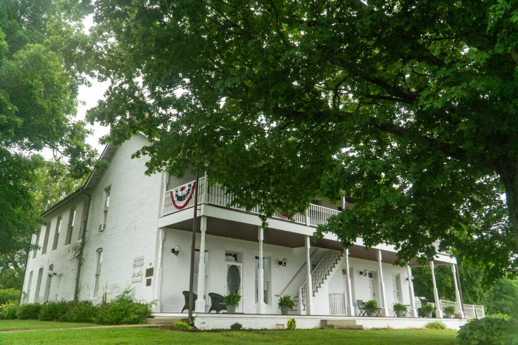 A white two-story house with a wrap-around porch and an exterior staircase, adorned with patriotic bunting. This is the Historic Rose Hotel, the yard is green with lush trees providing shade. Several chairs and potted plants decorate the porch, suggesting a welcoming, cozy atmosphere.