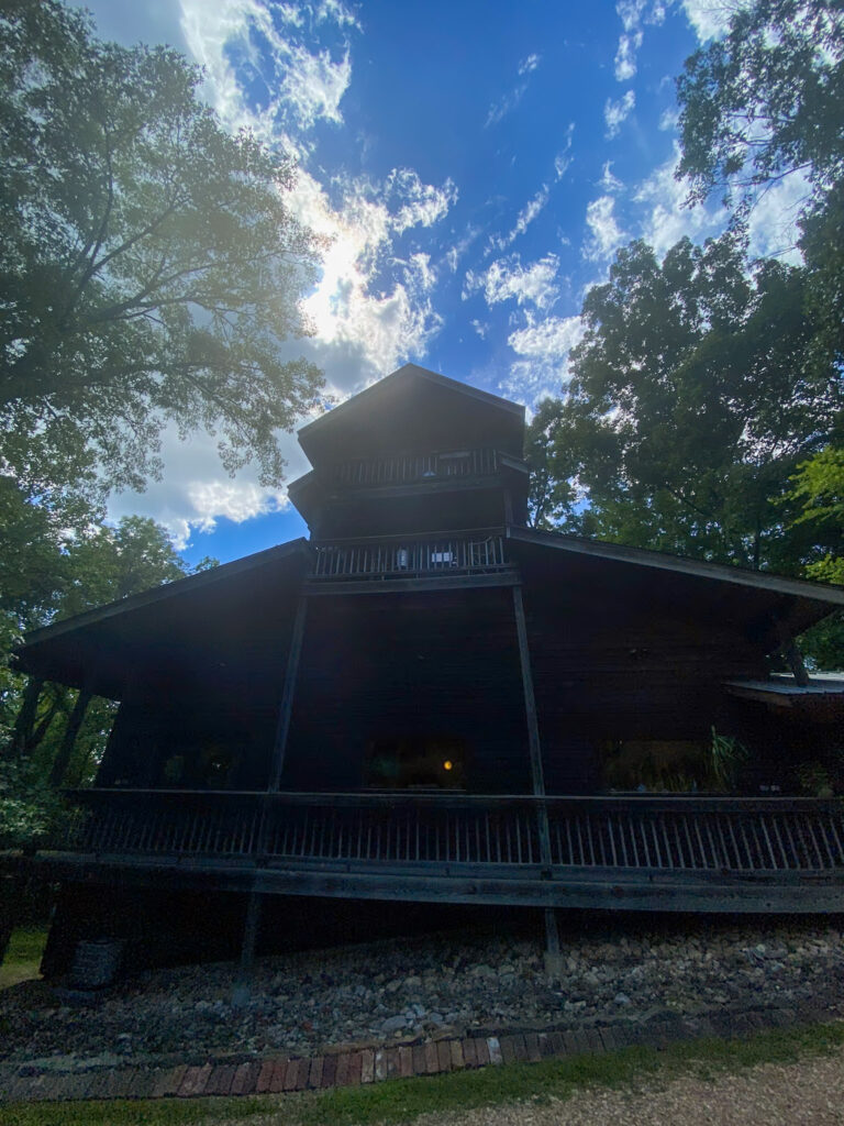 A dark, wooden cabin with a steep roof and multiple levels is surrounded by tall trees. The sun peeks through clouds in a bright blue sky above, casting a slight glow on the structure and creating dappled light across the scene.