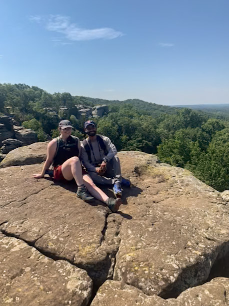 Two people are sitting on a large rocky outcrop with a lush, green forest stretching out behind them under a clear blue sky. Both are dressed in casual hiking attire, smiling, and appear to be enjoying the scenic view.