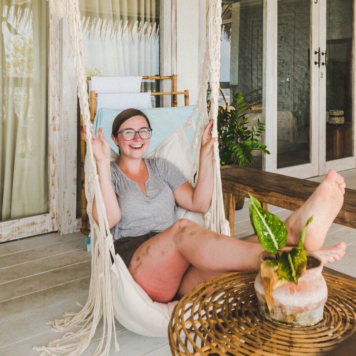 A person with glasses and a cheerful expression relaxes on a hanging chair on a porch. Their feet are propped up near a small table with a plant. The background features glass doors and a curtain.