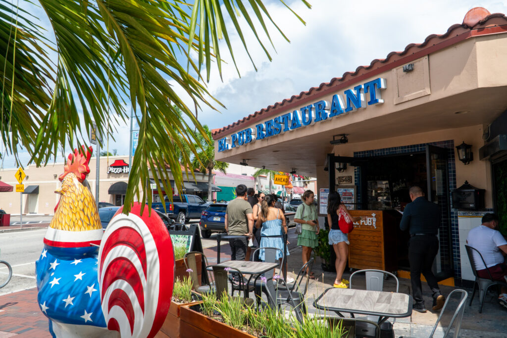 People gather outside El Pub restaurant with colorful signage, embarking on a Little Havana food tour. A large patriotic rooster statue stands in the foreground, as palm leaves partially frame the scene against a blue sky backdrop.