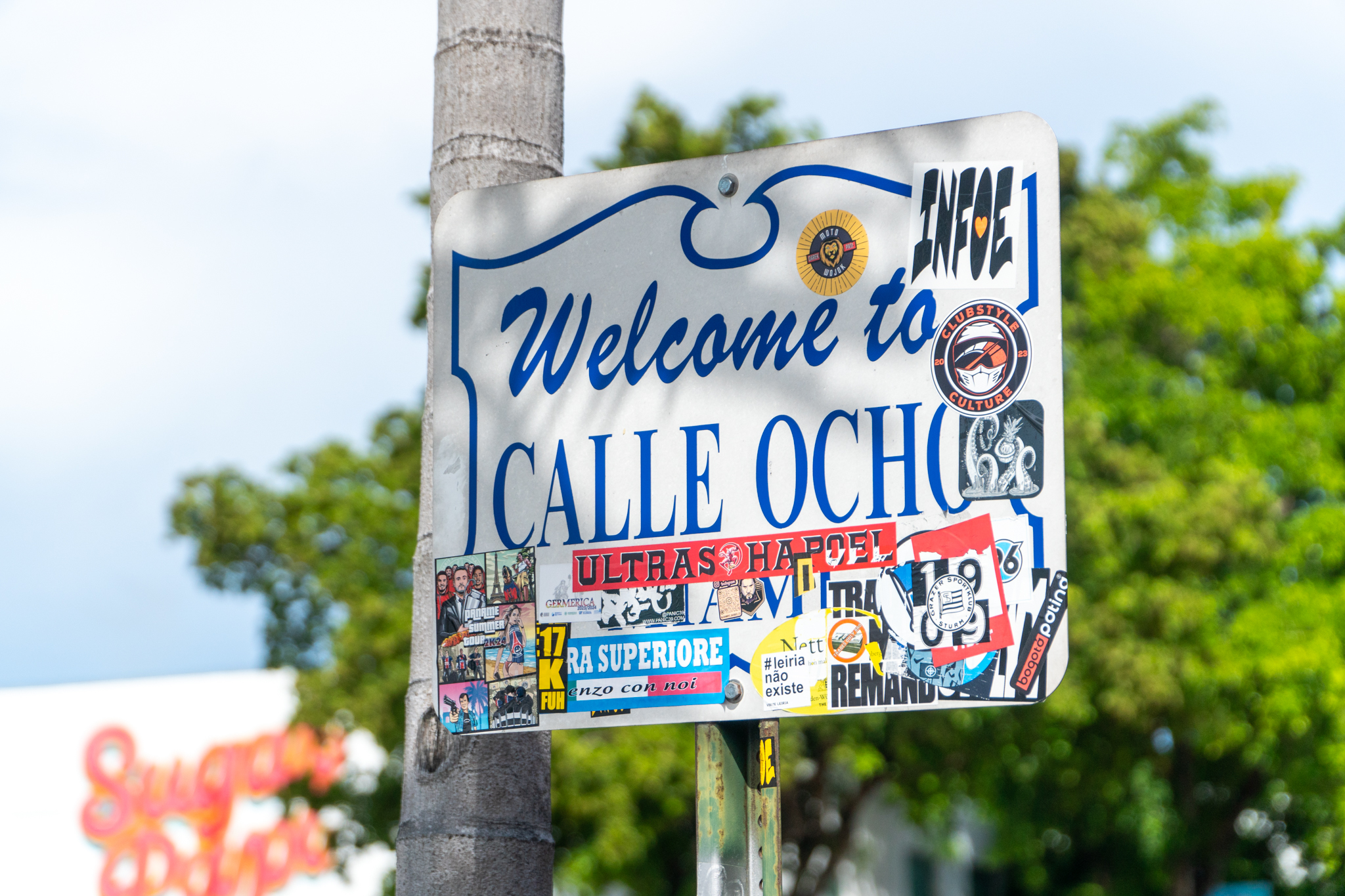 Sign reading "Welcome to Calle Ocho," covered with various colorful stickers, mounted on a pole with trees and a building in the background.