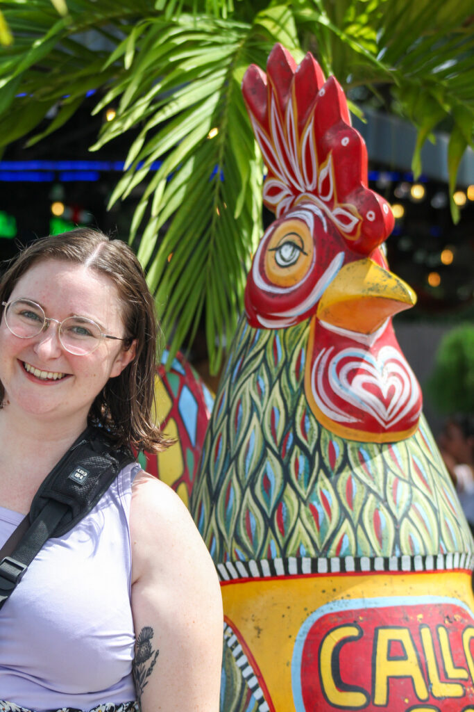 A person with glasses smiling next to a large, colorful rooster statue during a Little Havana food tour. The intricately patterned statue stands gracefully under palm leaves.